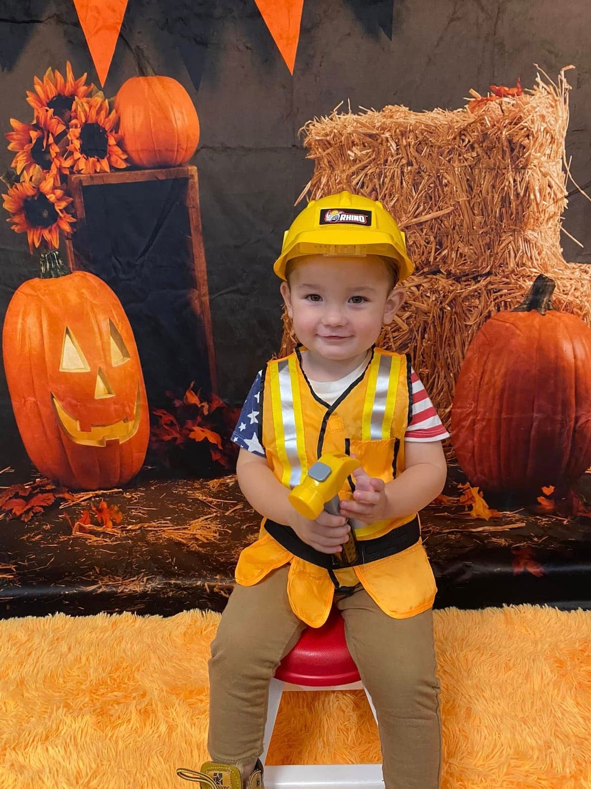 A toddler in a construction worker Halloween costume posing with pumpkins and hay bales