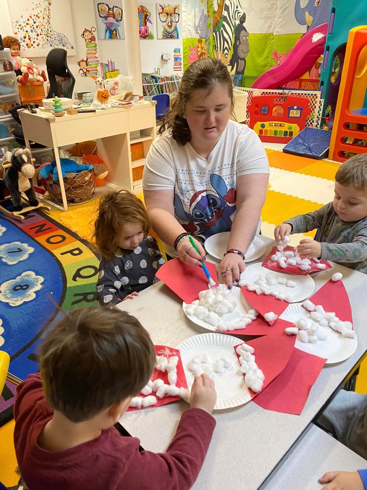 A teacher helping kids glue cotton balls onto Santa beard crafts at the playhouse table